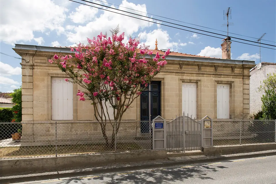Échoppe en pierre blonde à Saint-Médard-en-Jalles, avec volets blancs, clôture basse et lagerstroemia en fleurs devant la façade.